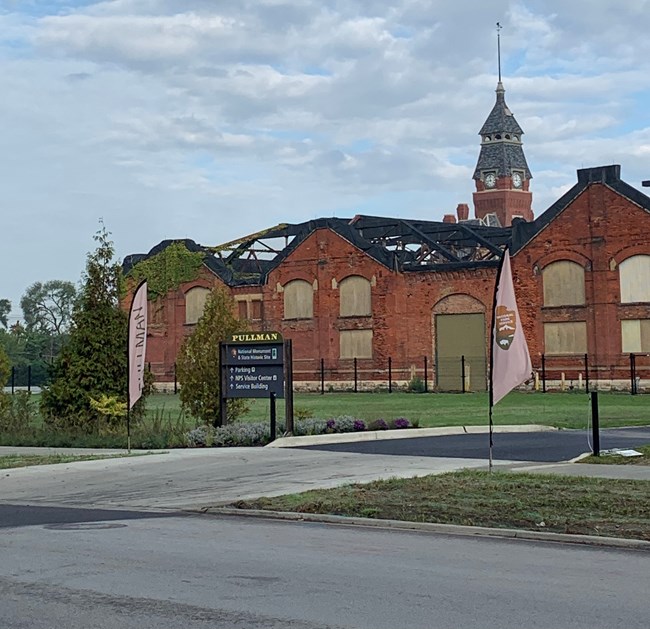 Parking Lot Entrance from street. Fin Flags flank either side of entrance. Old industrial red brick buildings in background.