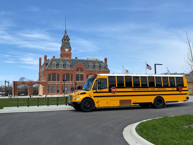 A yellow bus sits in the parking lot of the clock tower administration building on a partly cloudy blue-sky day.