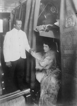 Photo of an African-American Pullman Porter serving a drink to a seated woman.