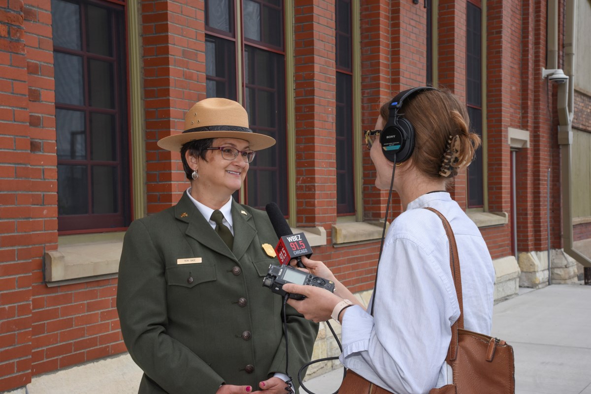 A park ranger in formal NPS green jacket and wearing a strawhat smiles at a reporter who holds up a mic and wears headphones.