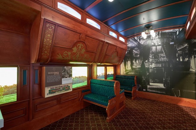 scenic railcar exhibit The inside of a replica train car with decorated wooden overhead bunk storage and cushioned green seats.