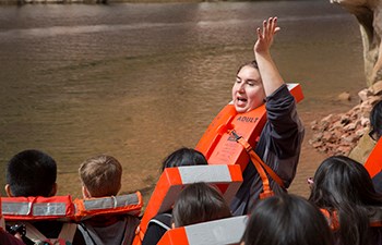 Woman wearing life jacket raises her hand while speaking to group of kids also in lifejackets