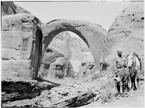 Historic photo of a man and a horse posing in front of Rainbow Bridge. First photograph of Rainbow Bridge taken.