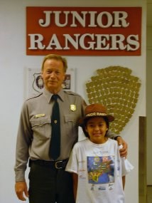 A Park Ranger stands with a child in a ranger hat in front of a sign that says Junior Rangers. A Park Ranger stands with a child in a ranger hat in front of a sign that says Junior Rangers.