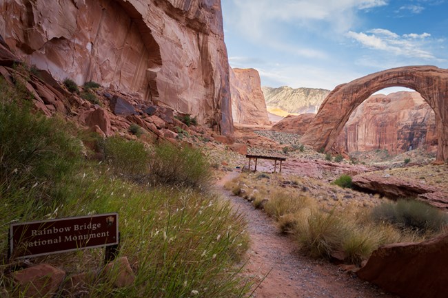 A large stone natural bridge stands surrounded by cliffs.  A sign in the foreground reads 'Rainbow Bridge National Monument.'