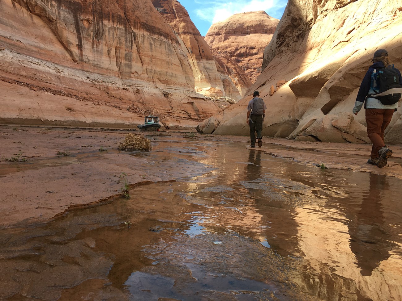 Two park rangers walk away from the camera towards a boat beached in a trickle of water in deep canyons.rds