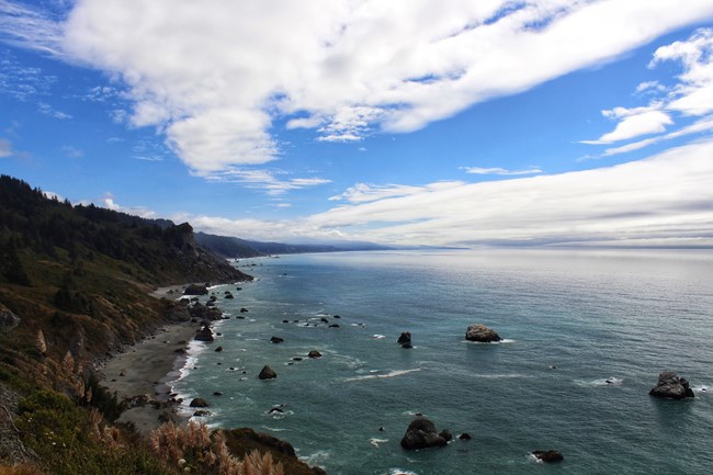 On a partially cloudy day,  green ocean and waves crash against tall, forested cliffs.