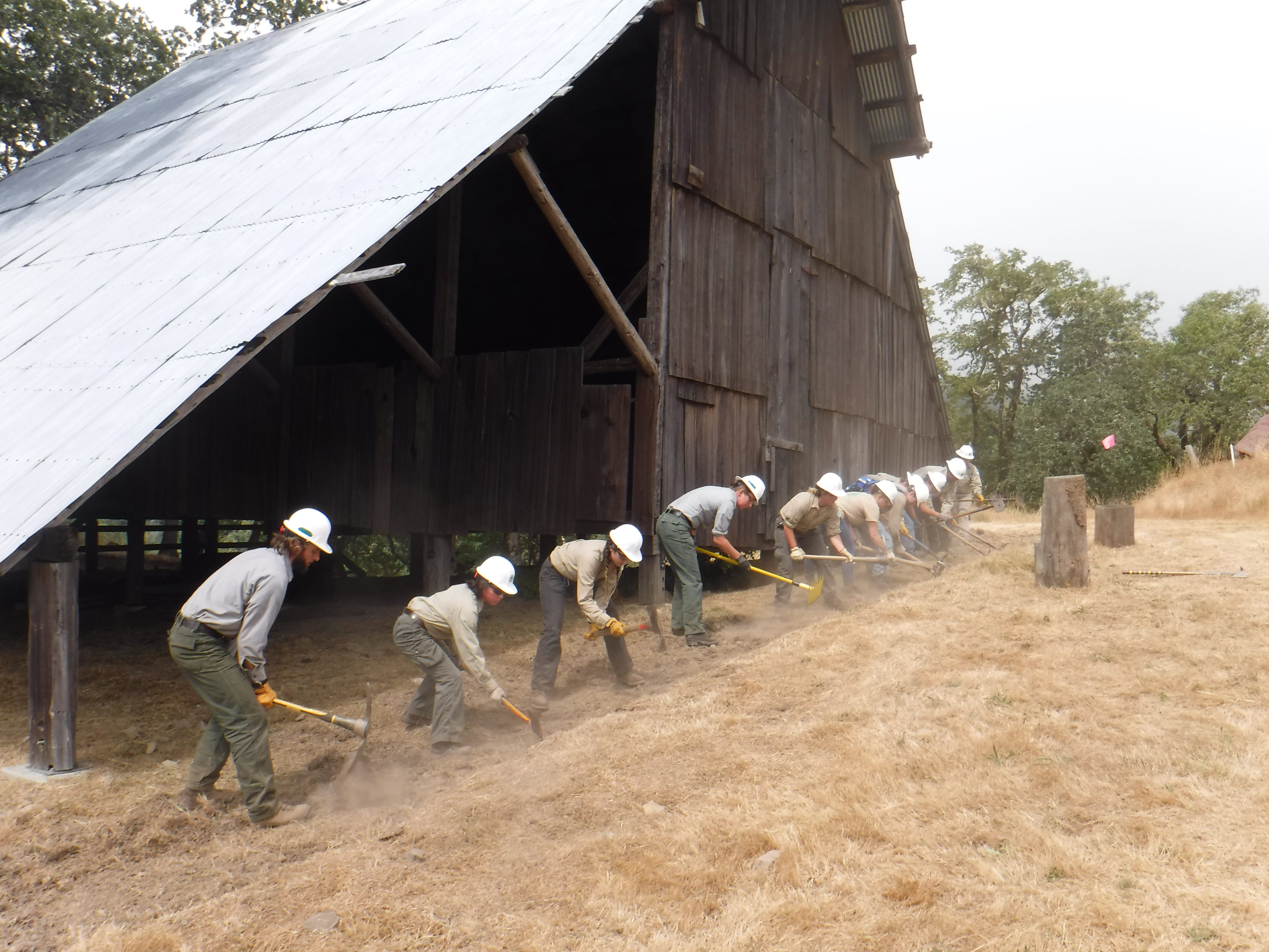 A dozen young adults use hand tools next to a historic barn