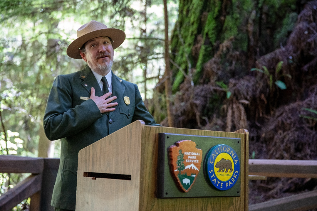 A middle-aged white park ranger stands on a podium next to a redwood tree.