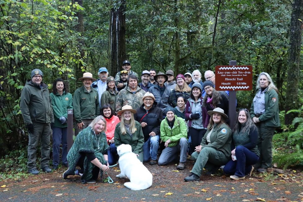 A large group of adults and a dog gather at a trailhead.