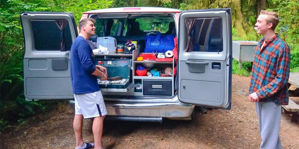 Two men cook breakfast by their camper-van.