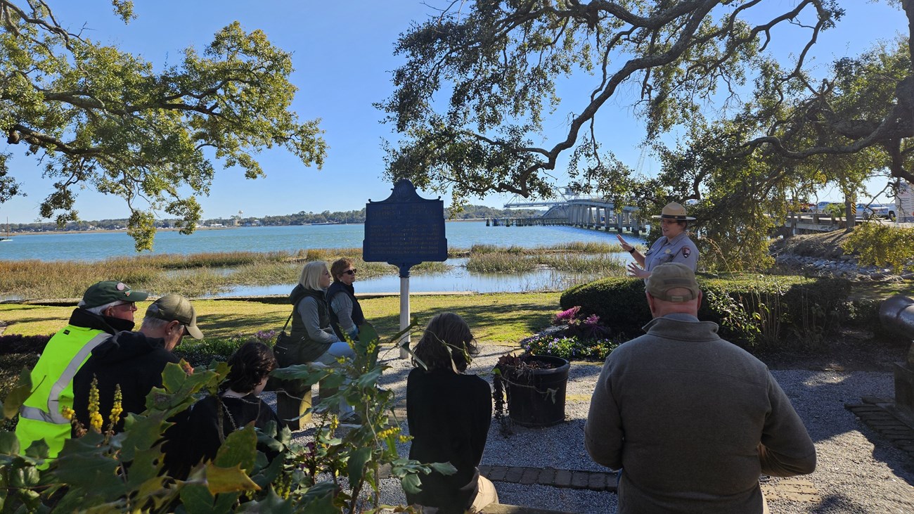 A group of people sit under trees in front of a marsh view listening to a ranger talk.