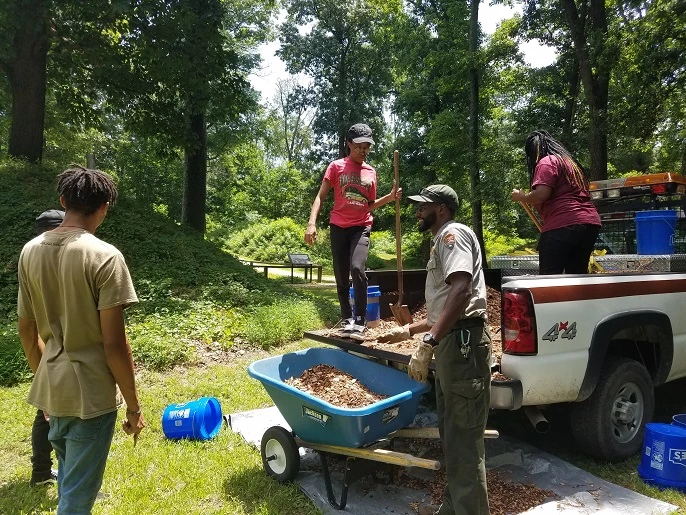 A park ranger with youth workers shoveling earth from the bed of a pickup truck.