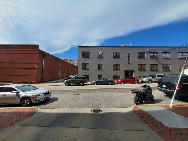 a three-story gray brick building on city street lined with parked cars