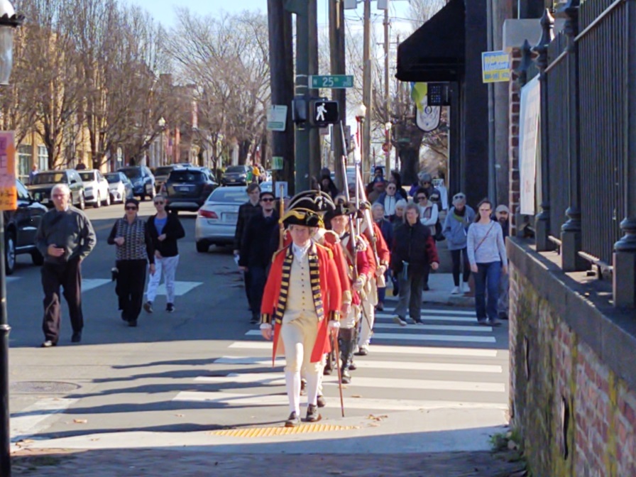 Benedict Arnold's Raid on Richmond Costumed reenactors in red Revolutionary War uniforms march in a line down a modern city street.