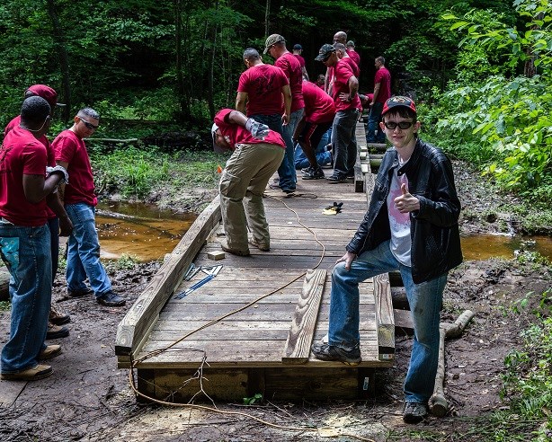 More than a dozen students in red shirts constructing a wooden bridge across a creek.