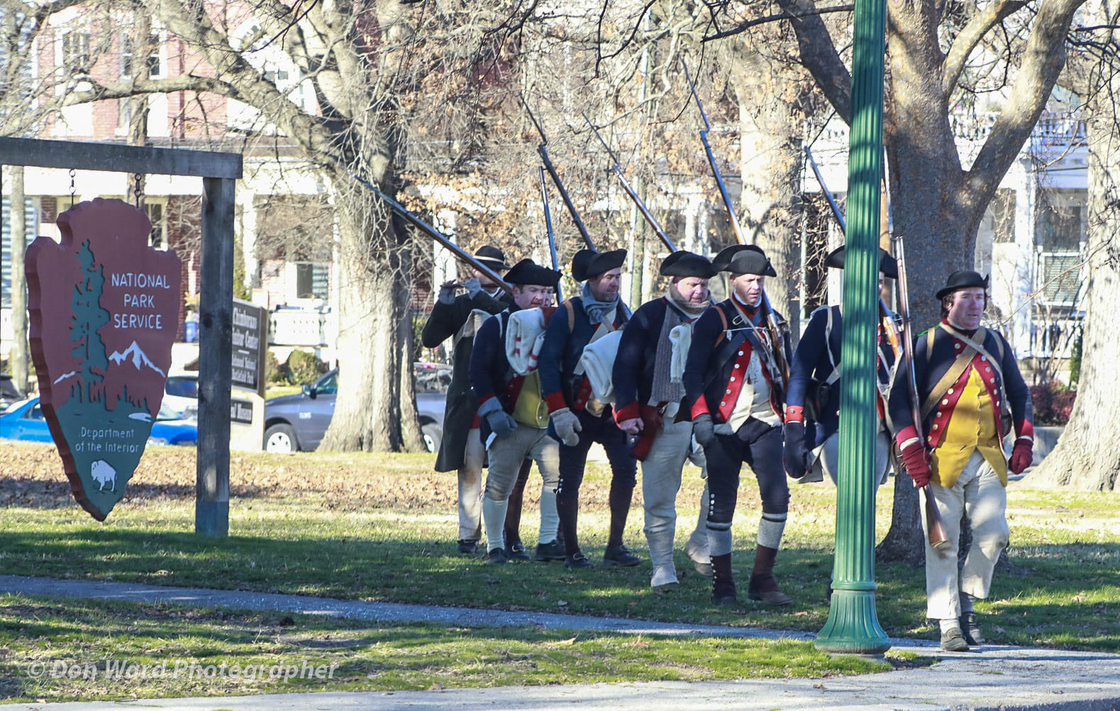 Benedict Arnolds Raid on Richmond Seven men in Revolutionary War attire march past a large, wooden arrowhead sign reading "National Park Service"