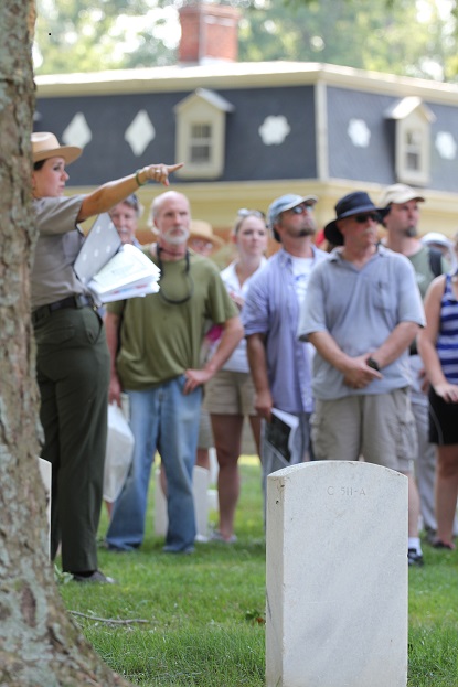 A uniformed park ranger gestures while standing among gravestones and park visitors