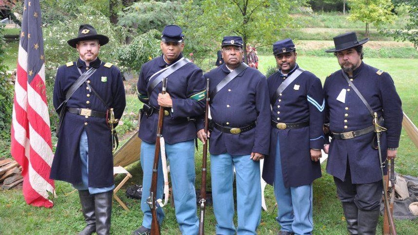 Black and white reenactors dressed in blue Civil War uniforms standing beside a large American flag.