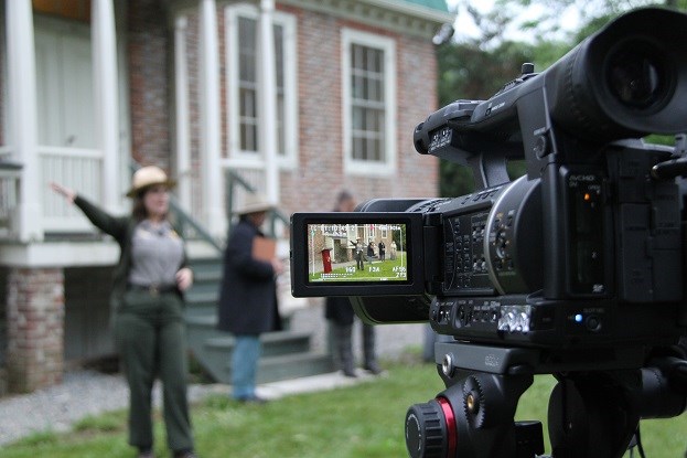 A park ranger, speaking, is visible in the viewfinder screen of a digital movie camera on a tripod