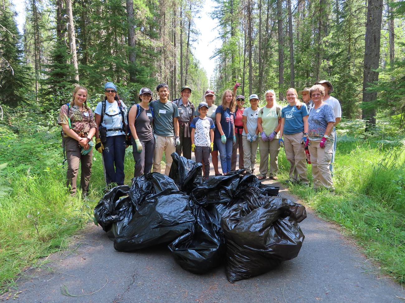 A group of proud volunteers stands in a forest behind garbage bags of weeds pulled during Glacier National Park's 2022 Noxious Weed Blitz