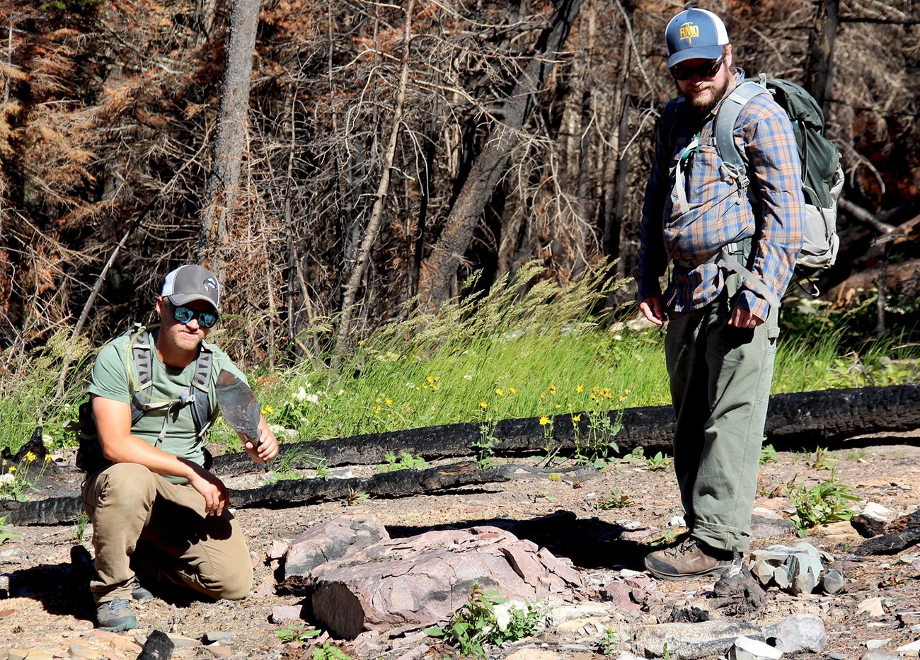 Two archeologists survey remains of a historic trapper cabin. A crouched young man holds an artifact while another archeologist stands surveying site.