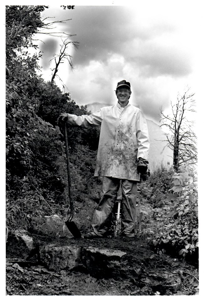 Man in rain gear stands on trail holding a shovel.