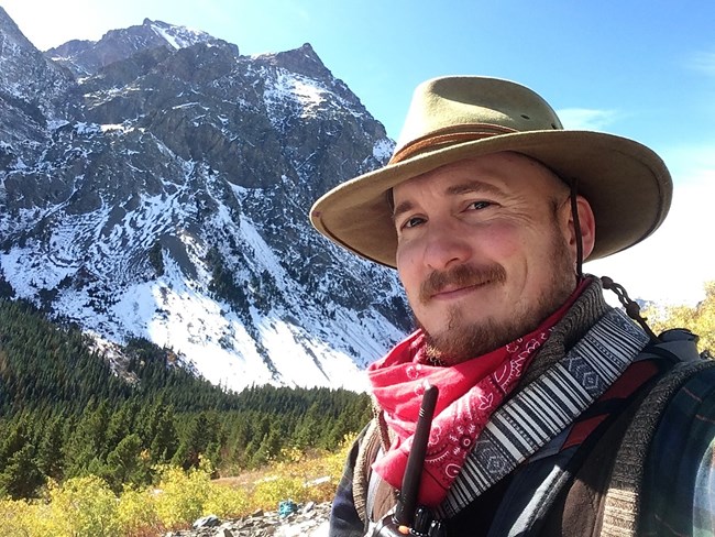 Close-up shot of Erik Nelson wearing a red bandanna and an expedition hat. A snow-covered mountain looms in the background.