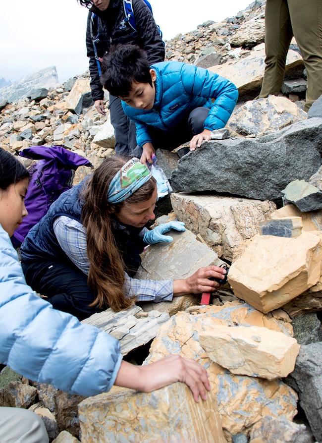 A woman holds a light to peer under a rock in a talus slope. Two kids look on.