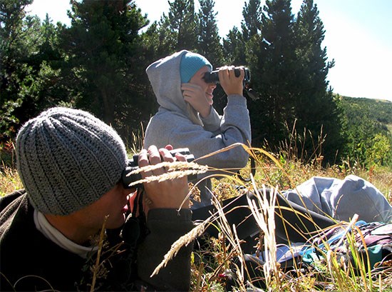 High high school students conduct a citizen science survey. Two high school students sit in meadow looking through binoculars.