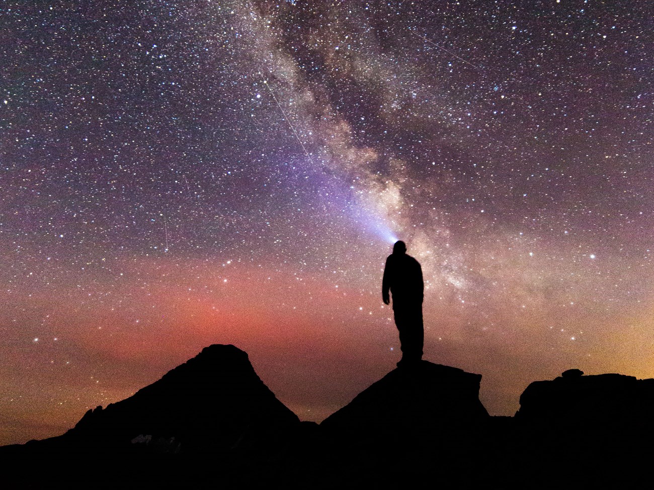 A hiker standing on rocks is silhouetted in black while looking up at the Milky Way at night
