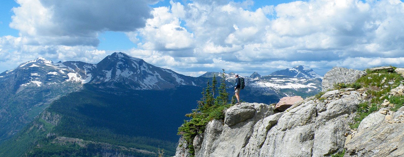 Hiker stands on rock cliff with mountain range in background.