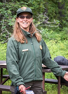A woman in ranger uniform and sunglasses stands in front of picnic table.