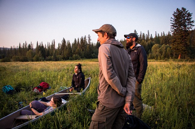 Four people wait on the shore of a small lake. Two of them sit in a canoe, and two others scan the lake with binoculars.