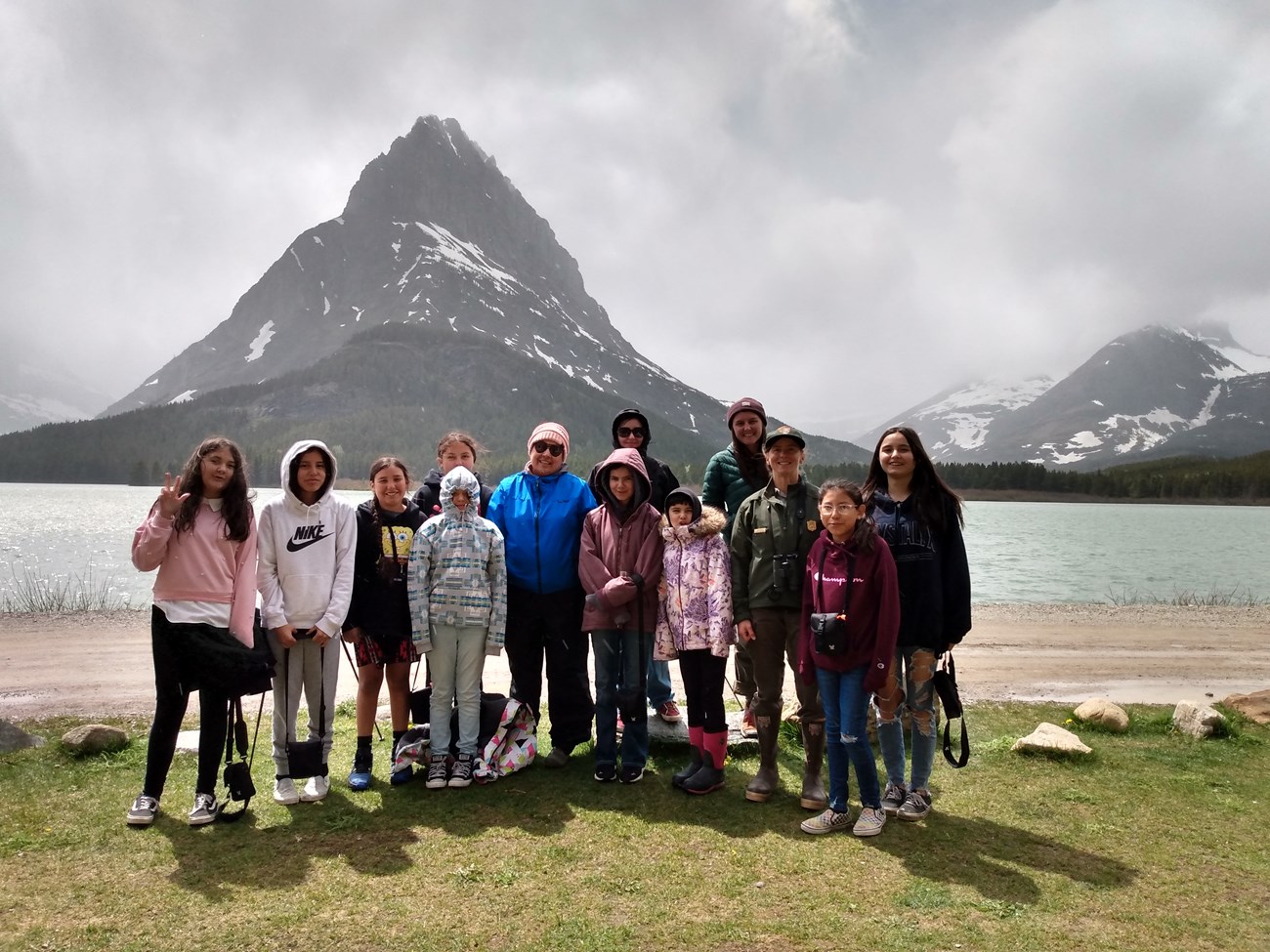 Nine young girls and four women (one in NPS uniform) stand on green grass in front of a light blue lake with a large gray conical mountain (Sinopah) and smaller mountains in the background.
