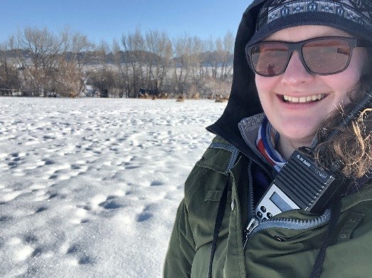 A woman has a radio tucked into her National Park Service uniform coat. She is standing in snow. Elk, trees, and hills are in the background.