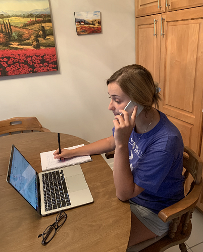 A woman sitting at a dining room table talks on her cell phone while looking at her computer screen and taking notes.