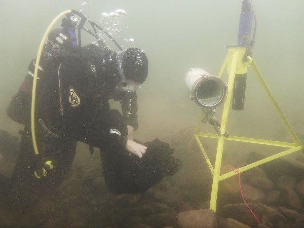 A SCUBA diver reaches into a bag for tools to service equipment clamped to a yellow tripod under the surface of Lake  Superior.