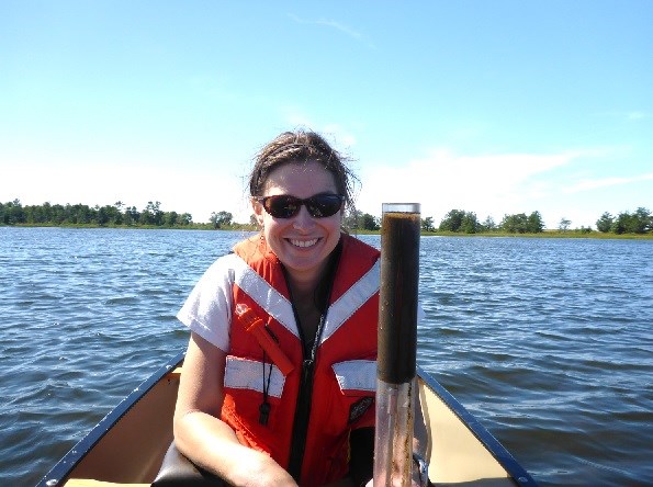 A woman in a PFD is in a canoe on the lagoon. She holds a clear cylinder partially full of sediment.