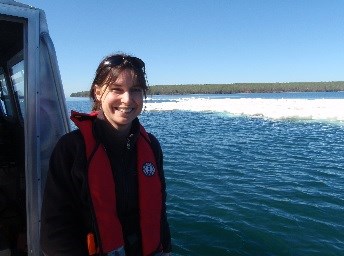 A woman stands on a boat in Lake Superior. Ice floats on the water and land is seen in the distance.