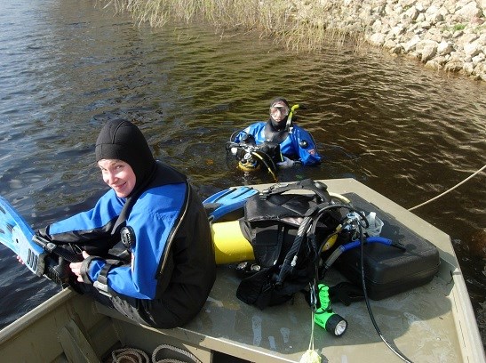 Assessments are conducted periodically to analyze changes in zebra mussel populations over time. A woman sits on the bow of a boat and puts on diving equipment. Another diver is in the water.