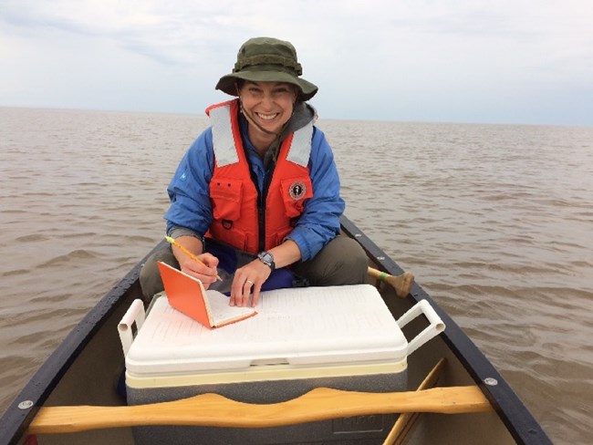 Viewed from the bow of a canoe in the open water, a woman sitting in the stern smiles as she writes in her notebook. She is using an ice chest as a table.