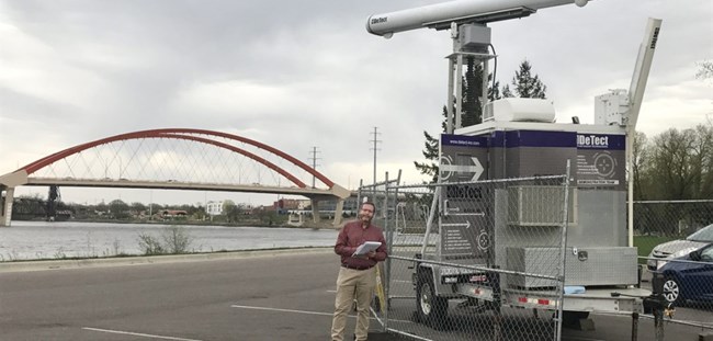 Research using the equipment shown here could help save the lives of birds in the future. A special trailer with avian radar monitoring equipment is set up behind a fence. The Clark Bridge appears in the background.