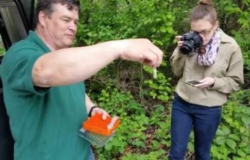 A young woman photographs a man holding a small plastic sample vial.