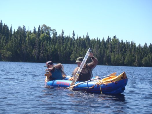 Two field technicians lower instruments over the edge of an inflatable boat and into the water.