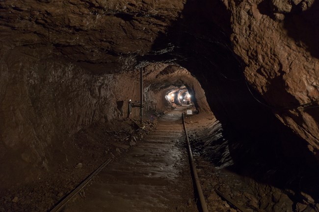 An underground view down a mine tunnel. Rails lead toward electrical lights in the distance.