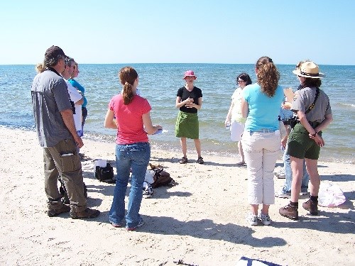 Eleven people are standing in a circle on a beach.  One person is dressed in park ranger uniform. Lake Michigan is in the background.