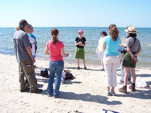 Eleven people are standing in a circle on a beach.  One person is dressed in park ranger uniform. Lake Michigan is in the background.