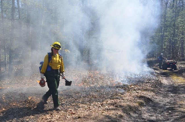 A woman in protective fire gear carries a drip torch. Smoke rises from a fire behind her.