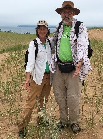 A woman and man stand behind a silvery stemmed thistle  on a sand dune. A large lake is in the background.